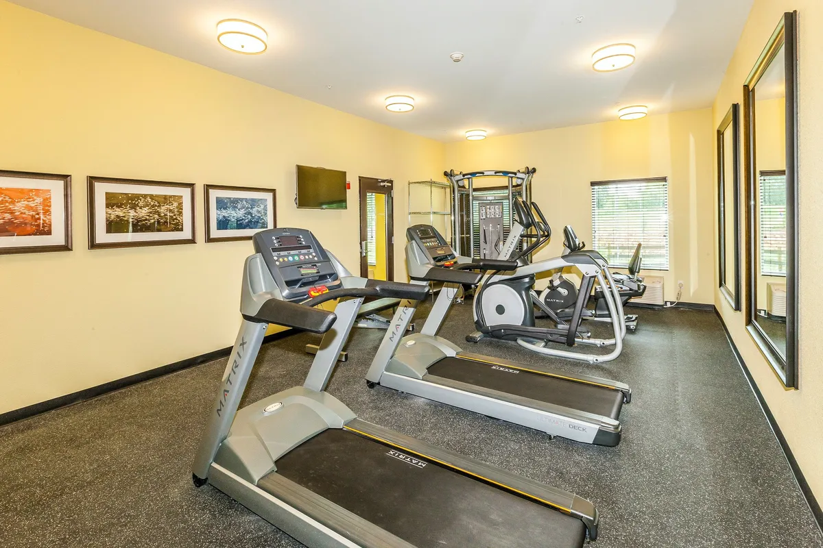 This interior shot highlights the modern 24-hour fitness center, featuring treadmills, an elliptical machine, weight training equipment, and wall mirrors. The atmosphere is bright and motivating, with gym-goers engaging in both cardio and strength training activities. Artwork on the walls and TVs create an energizing, contemporary feel.