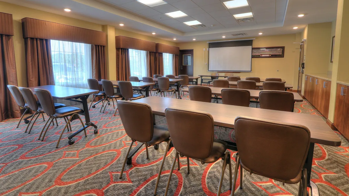 This image showcases an interior space set up for meetings, with tables and chairs arranged to foster collaboration. The large windows let in abundant natural light, creating a bright and welcoming ambiance. The space includes a prominent projector screen, suggesting a venue that can adapt to various functions, including casual gatherings or working meals.