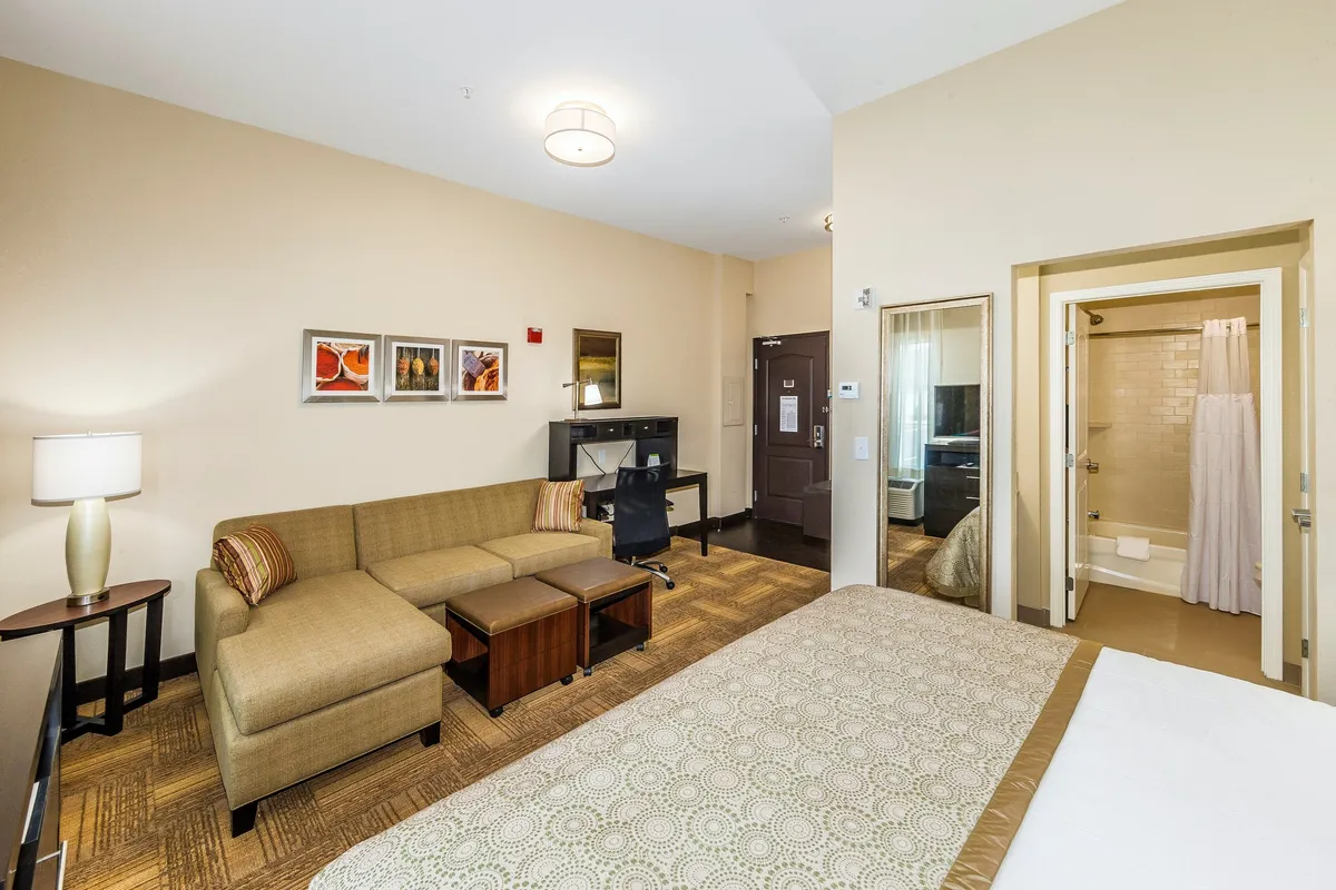 The image displays a spacious, modern studio suite featuring a plush bed, cozy sofa, practical desk, and a sleek shower area, all accented by tasteful artwork. The layout seamlessly blends relaxation, work, and leisure zones, with inviting lighting and thoughtful decor that create a welcoming atmosphere.