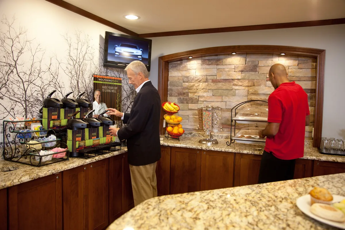 A well-appointed breakfast area with a modern coffee station, a fresh fruit display, tempting pastries, and cheerful interaction between a staff member and a guest. The mood is relaxed and friendly, ideal for starting the day with a meal or coffee.