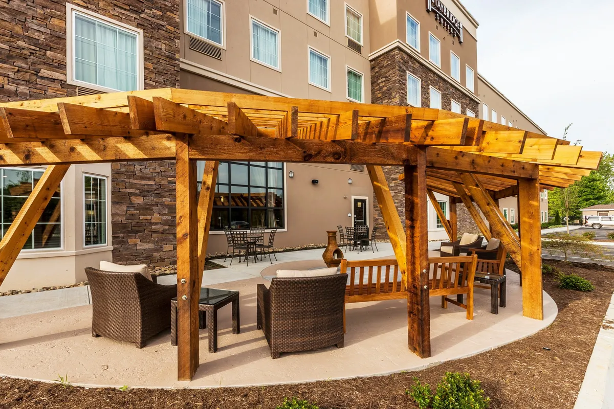 This image features a comfortable outdoor seating area under a wooden pergola, accompanied by a stone wall. The scene is inviting, suggesting a place for relaxation or socializing, and hints at proximity to amenities where one might enjoy coffee or a meal with friends.