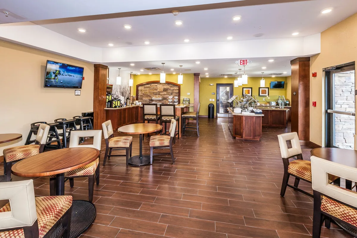 A spacious, welcoming dining area featuring circular wooden tables and upholstered chairs, with a stone feature wall and a breakfast bar displaying an assortment of food. The setup is bright and inviting, offering ample space for enjoying meals comfortably.