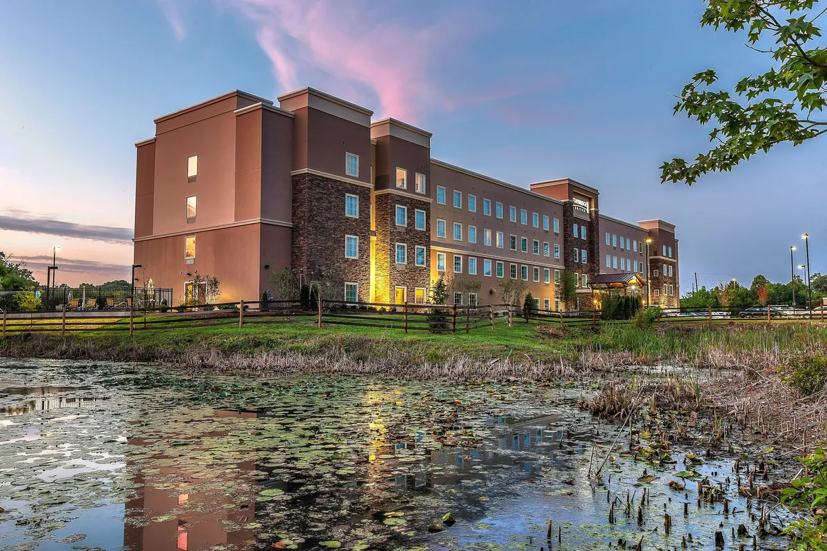 The exterior view highlights the hotel's facade reflected in water and surrounded by natural greenery. The scene is calm and inviting, suggesting opportunities for pleasant walks and relaxation within a lively yet scenic environment.